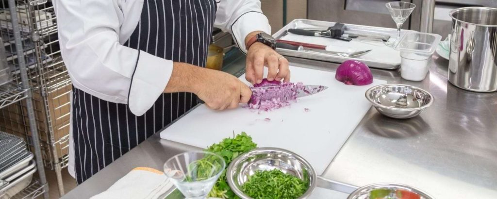Image of a student wearing a striped apron, chopping a red onion in a professional kitchen at Florida Education Institute